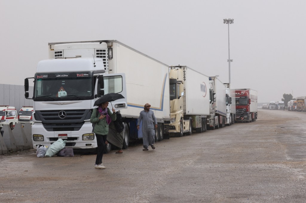 A reporter uses an umbrella to protect herself from the rain, as trucks transporting humanitarian aid wait at the Rafah border crossing between Egypt and the Gaza Strip, during a temporary truce between Hamas and Israel, in Rafah, Egypt, November 27, 2023. REUTERS/Amr Abdallah Dalsh